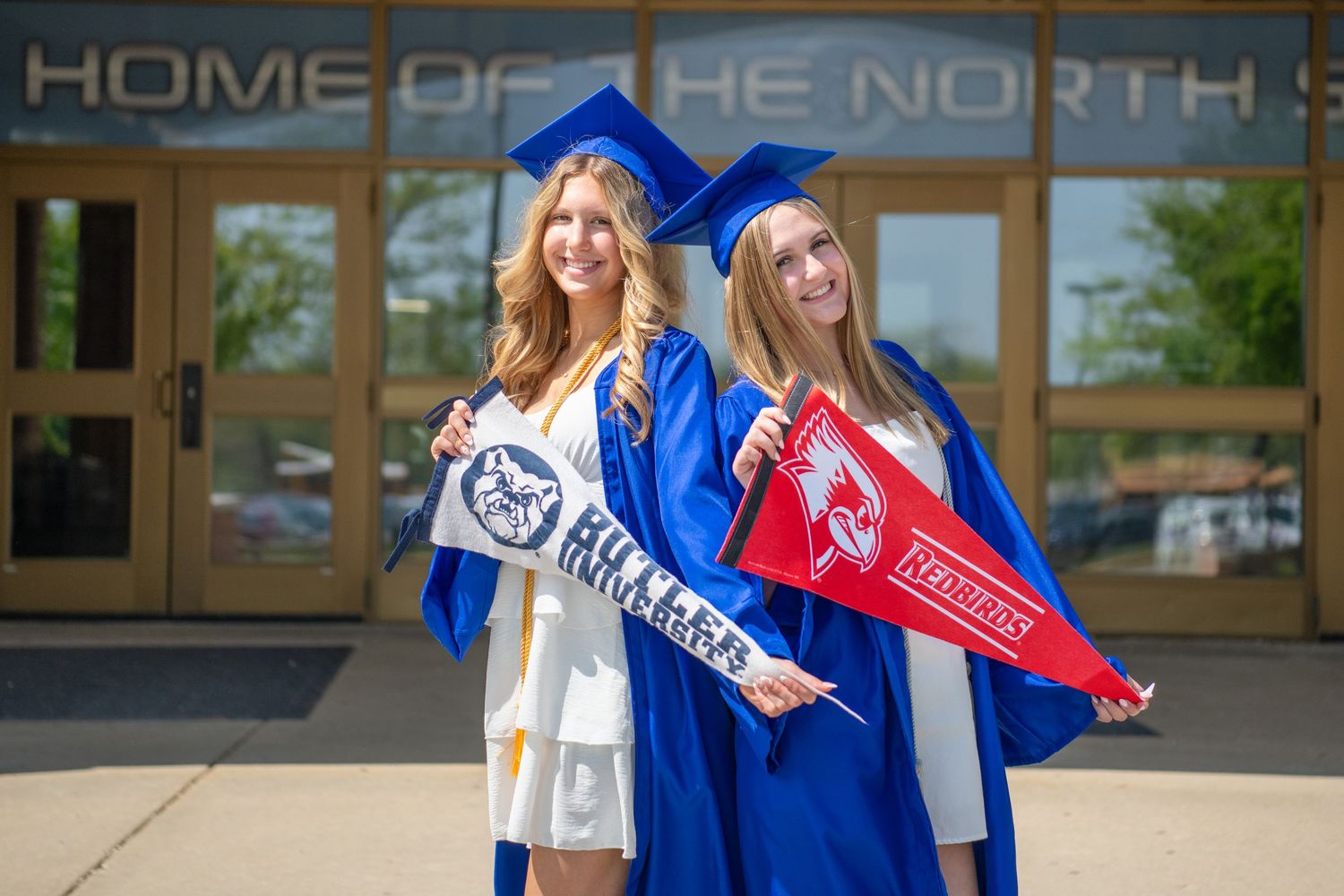 Two graduates in blue caps and gowns hold school pennants while standing outside a building entrance.