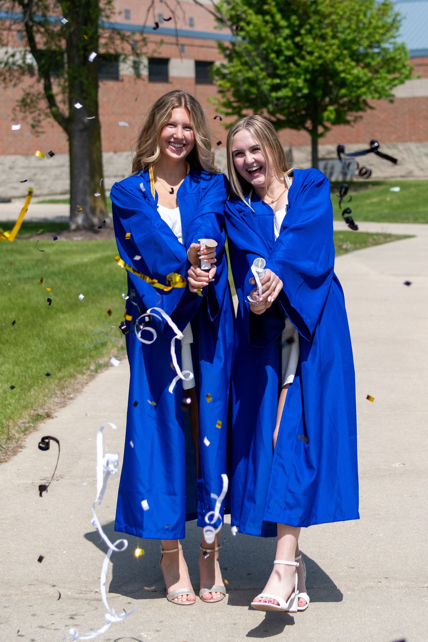 Two graduates in bright blue graduation robes celebrate on a sunny day outside a brick building.