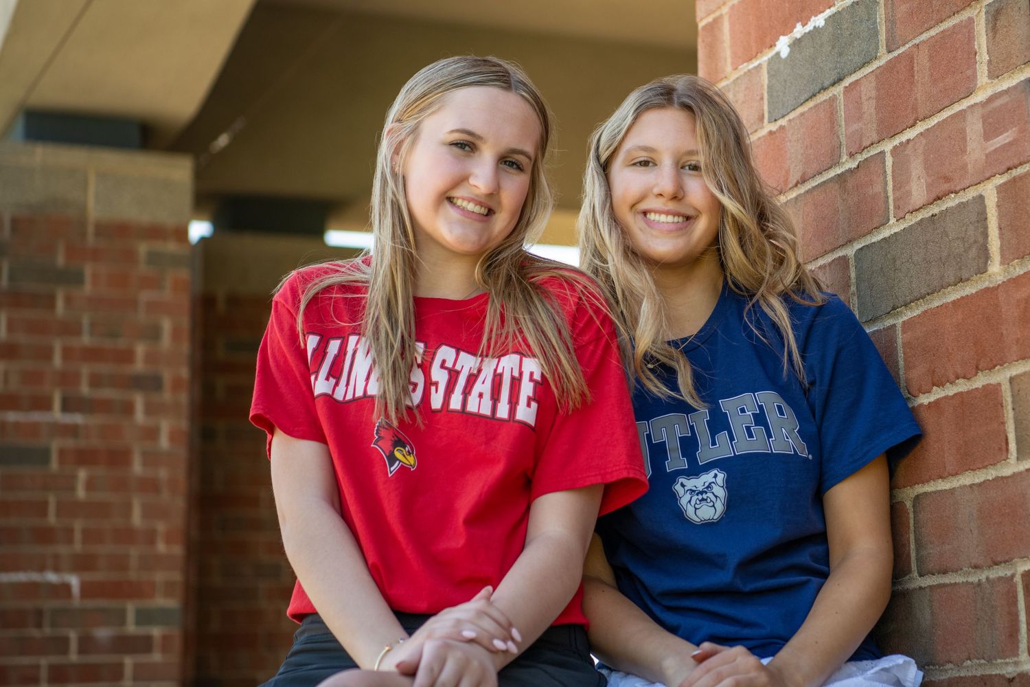 Two people in contrasting red and blue shirts sitting together on brick steps smiling at the camera.