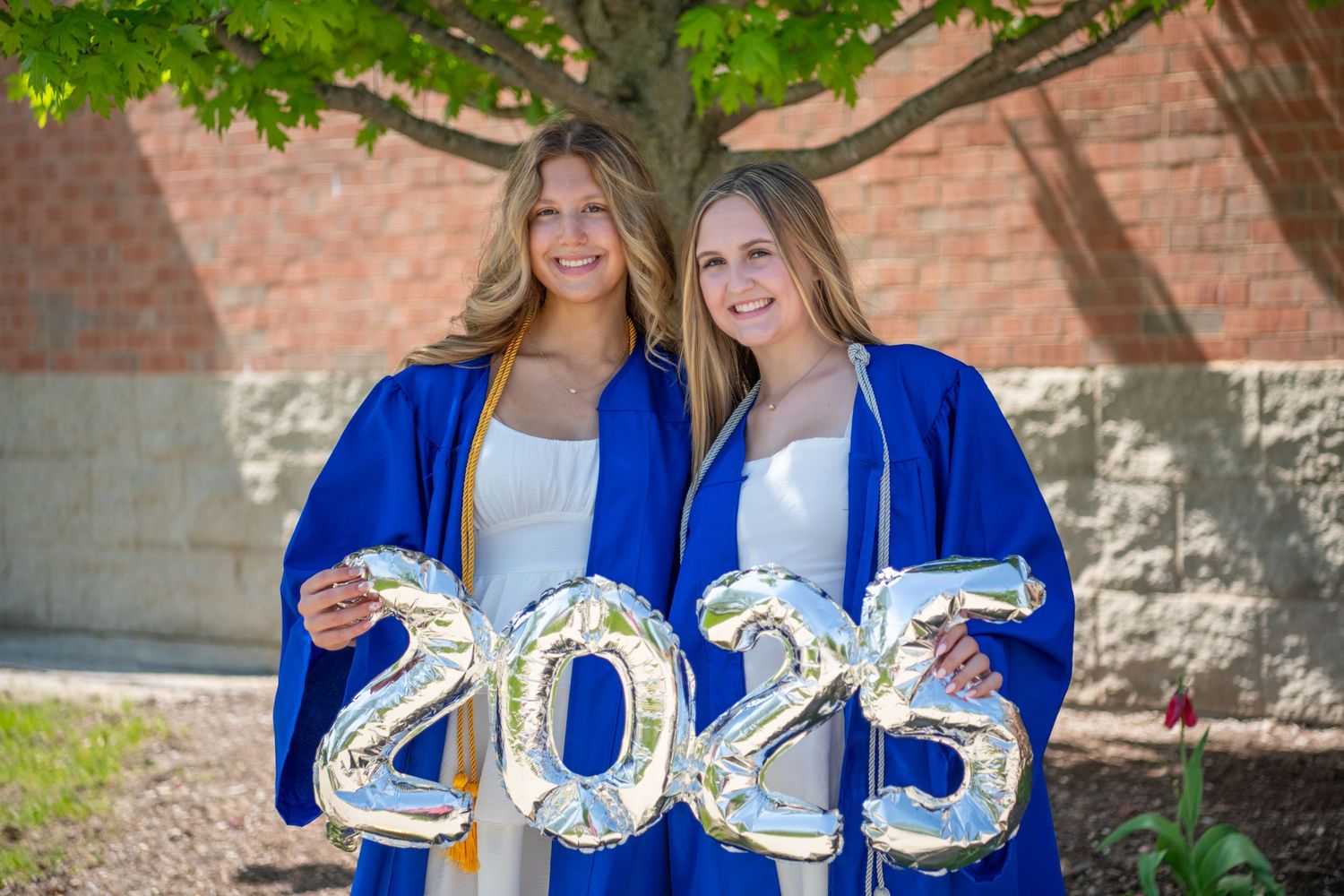 A group of people in royal blue graduation gowns hold silver balloon numbers outdoors near a brick building.