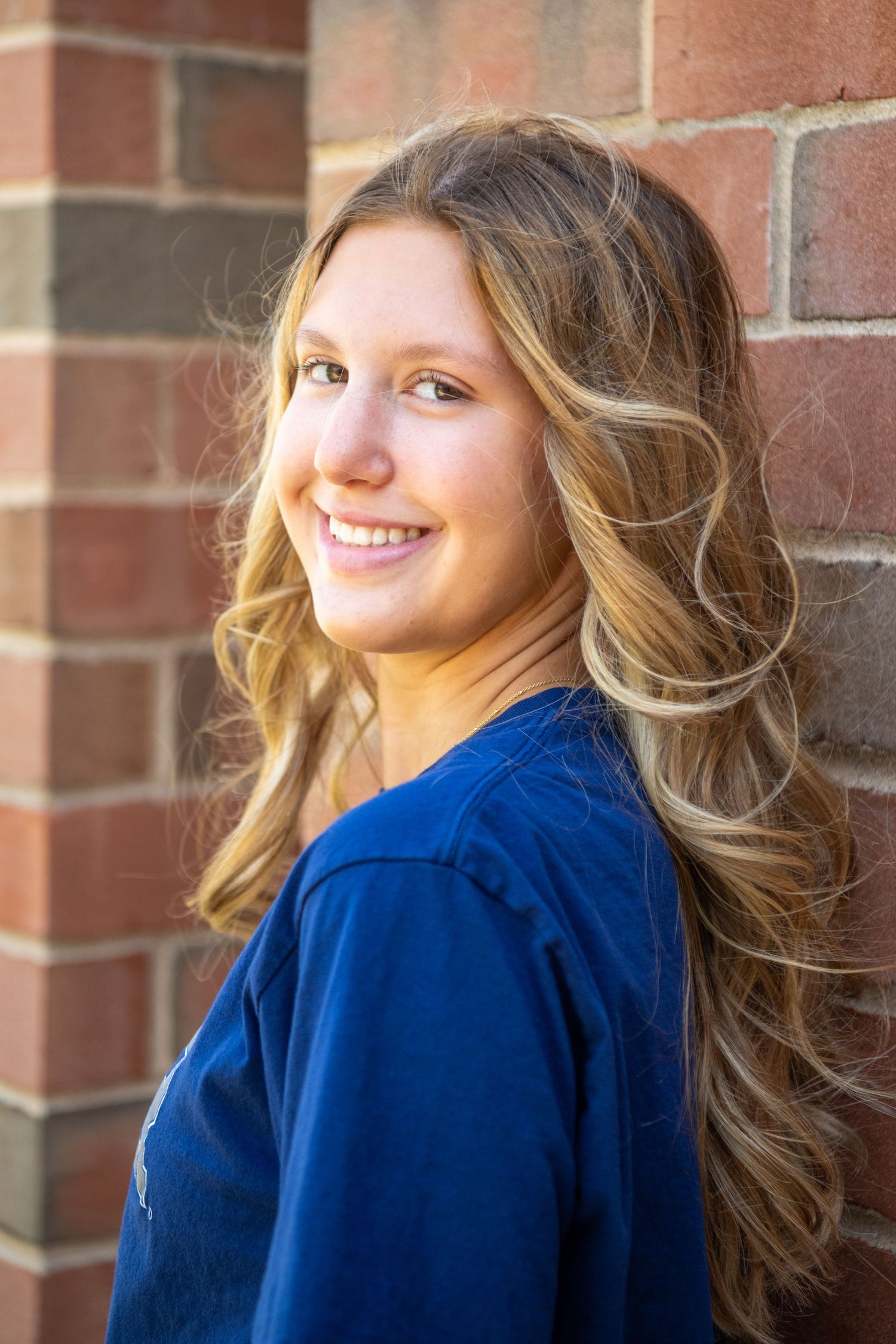 Portrait of someone with long wavy blonde hair wearing a navy blue top smiling against a brick wall background.