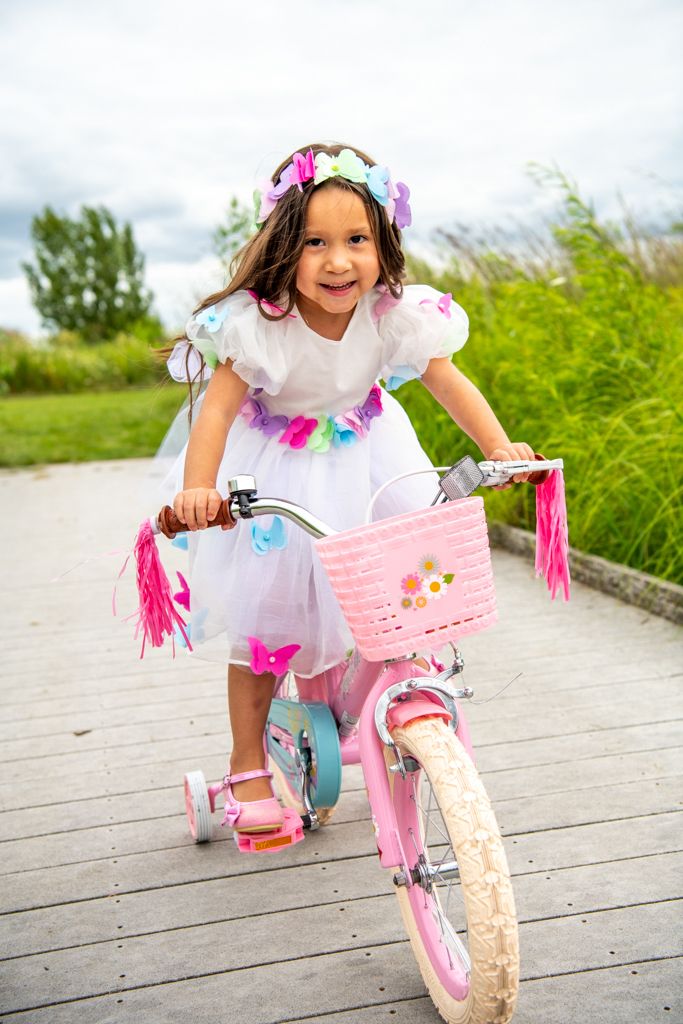 Young child in white dress with floral crown riding pink bicycle on wooden path.