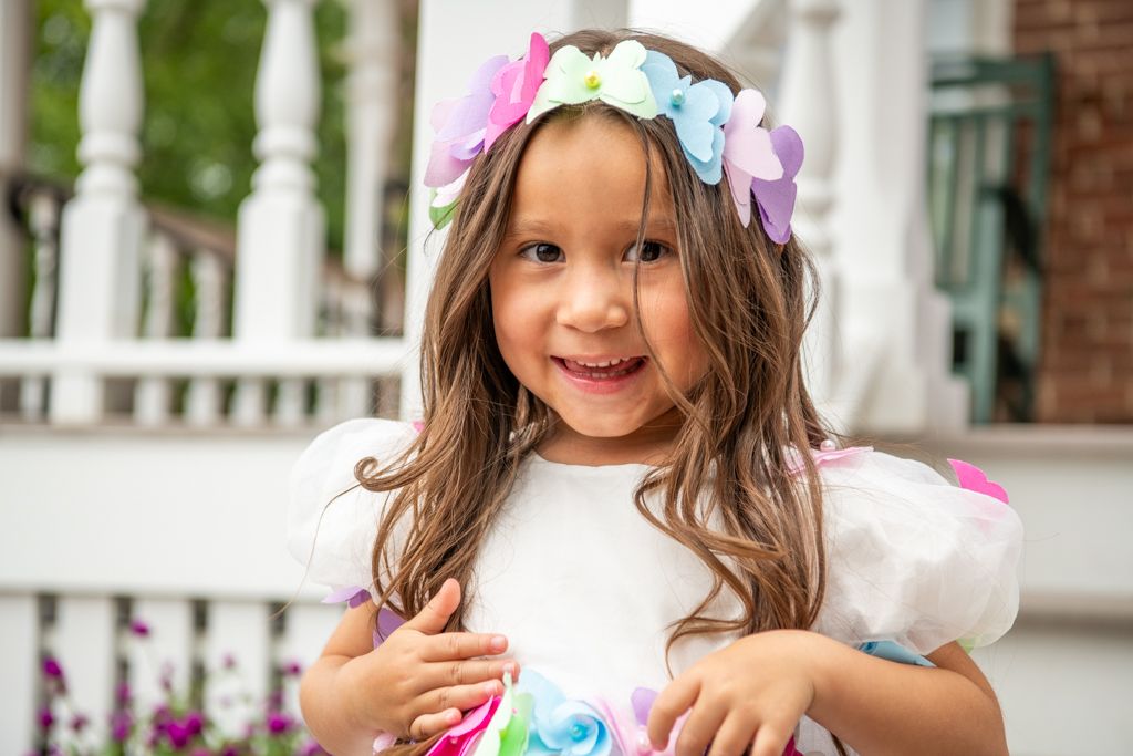 Young child wearing floral crown and white dress smiling on porch.