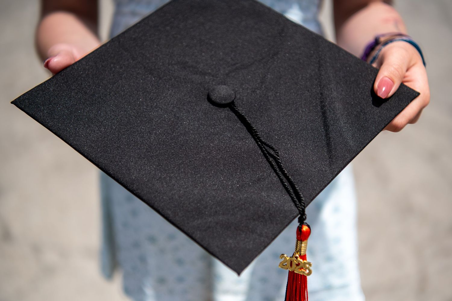 Black graduation cap with red and gold tassel held against blurred background.
