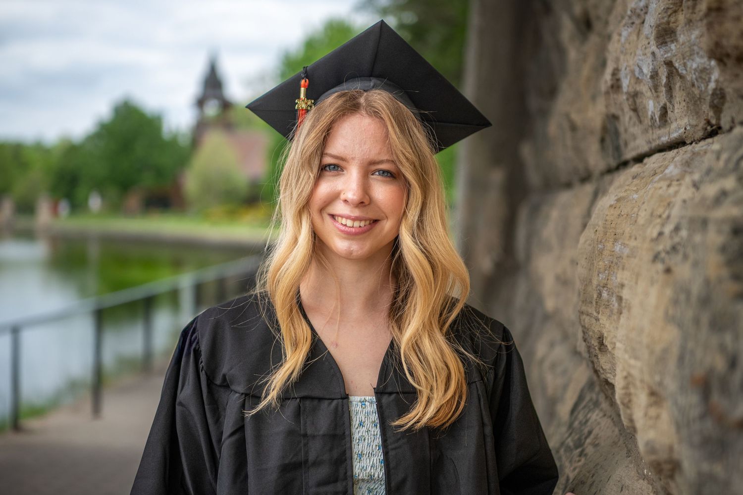 Graduate in black cap and gown poses against stone wall near waterfront.