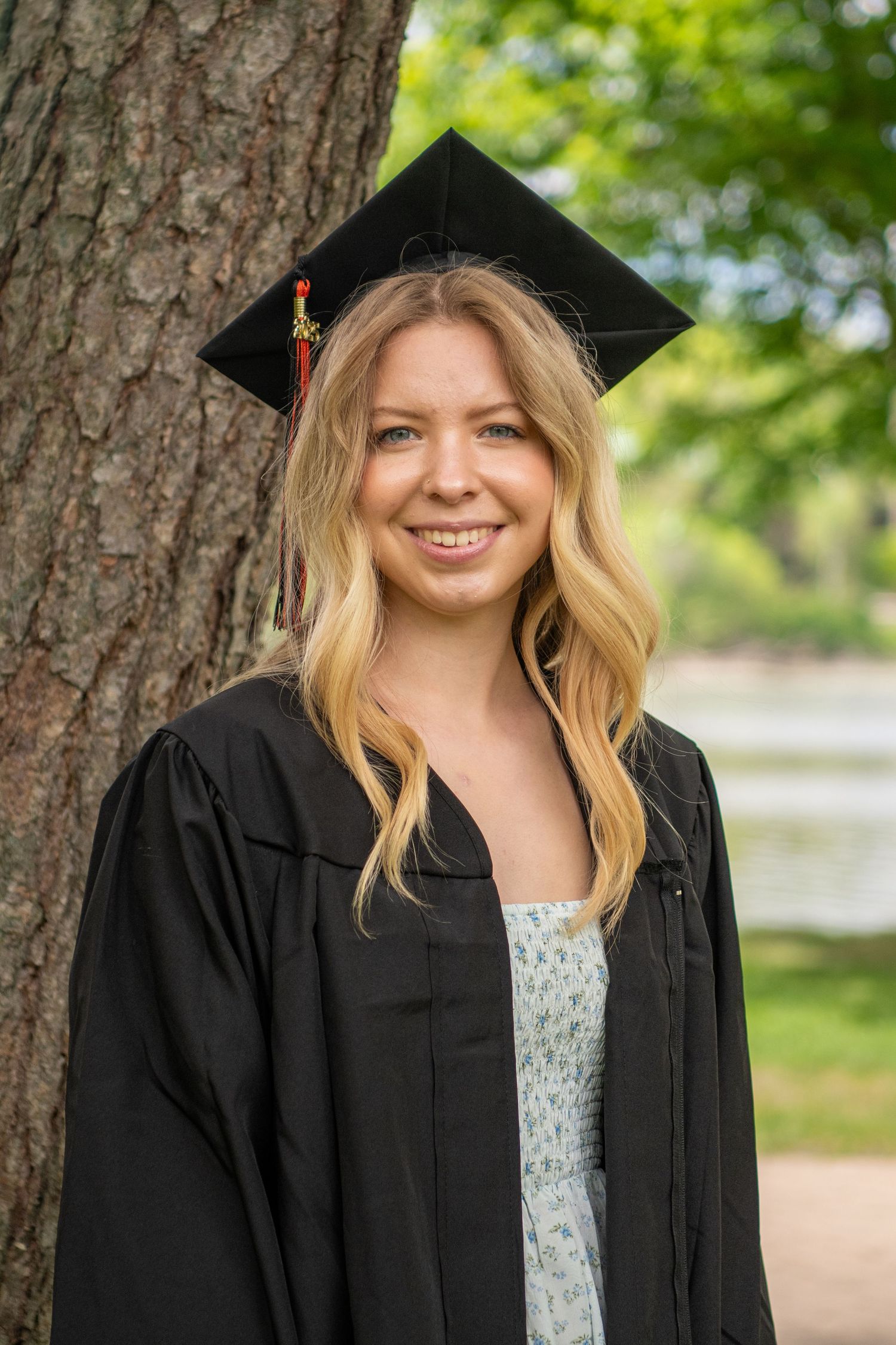 Graduate wearing black cap and gown stands in front of tree during outdoor photo session.