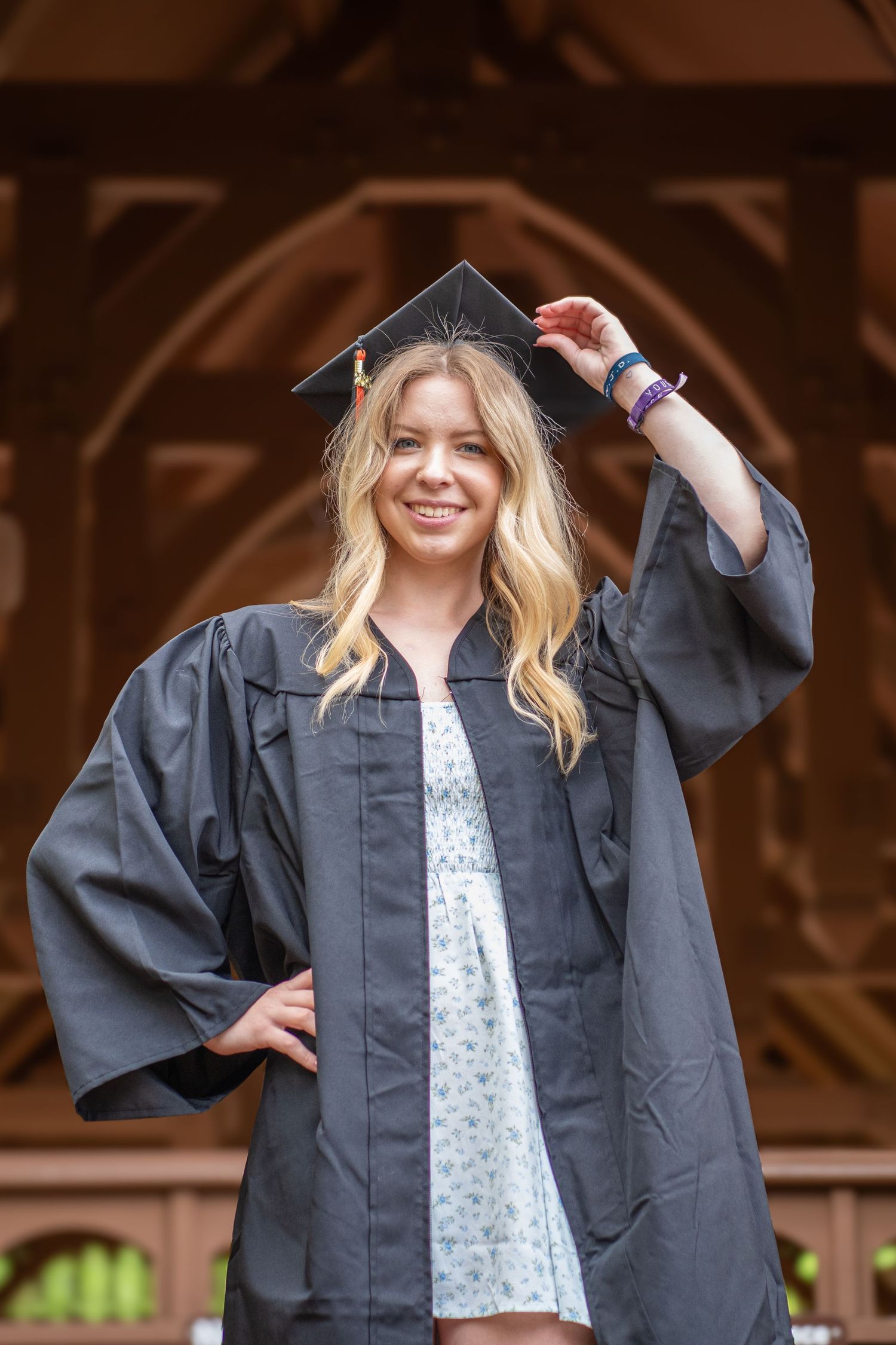 Graduate wearing traditional black graduation gown and cap poses outdoors in front of wooden architecture.