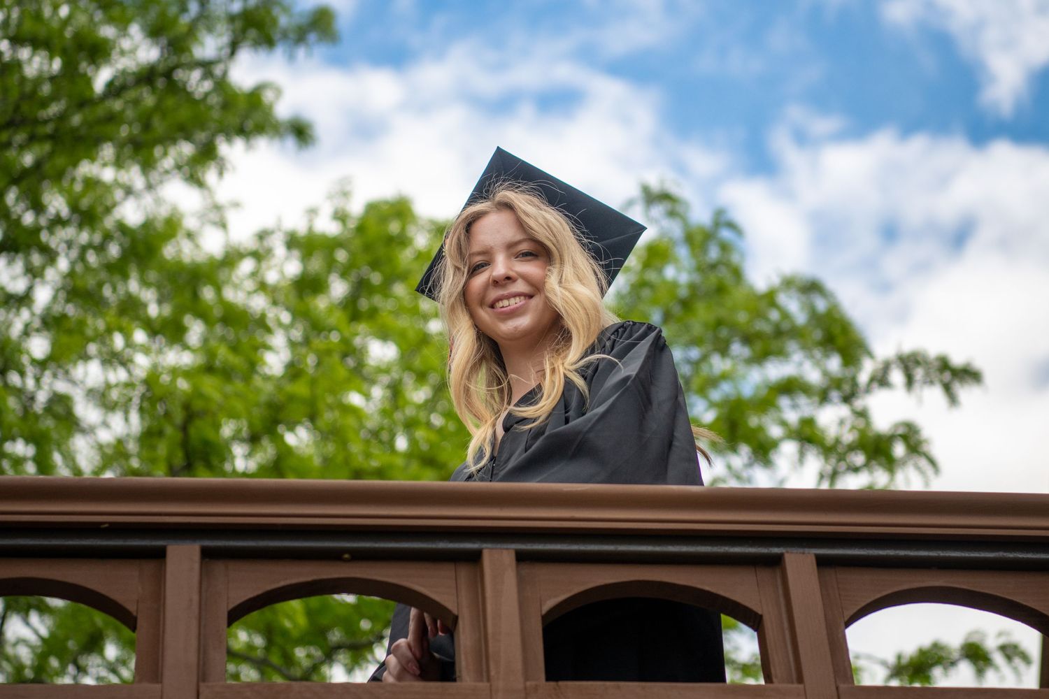 Graduate in traditional regalia stands on wooden bridge under blue sky.