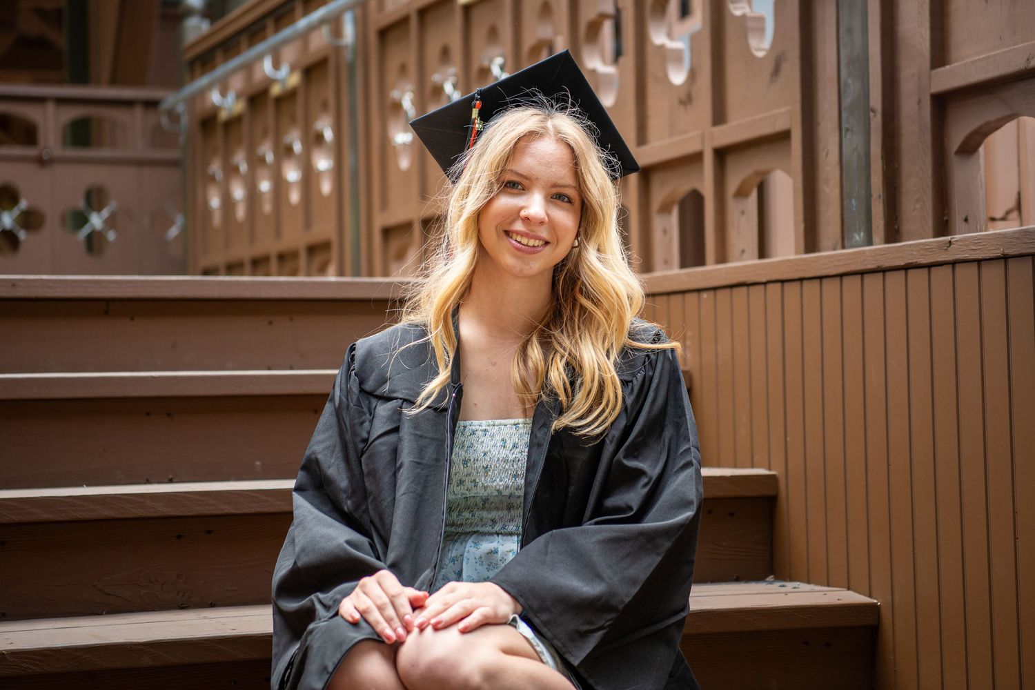 Graduate in denim jacket sits on wooden steps wearing cap and gown.