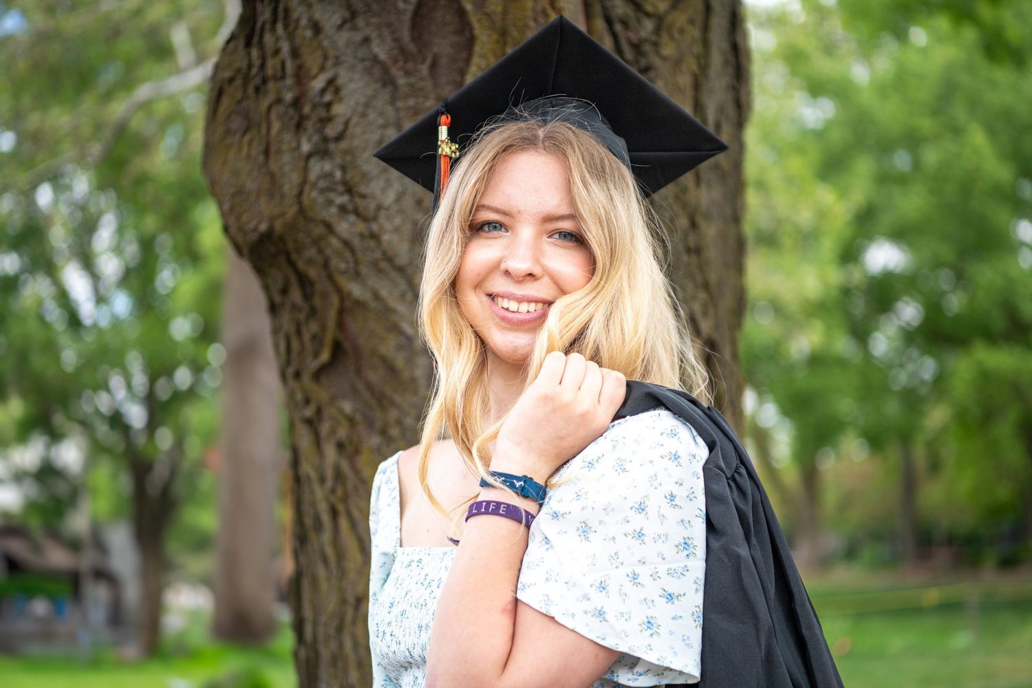 Graduate in black cap and gown poses outdoors in a park setting with trees and greenery in the background.