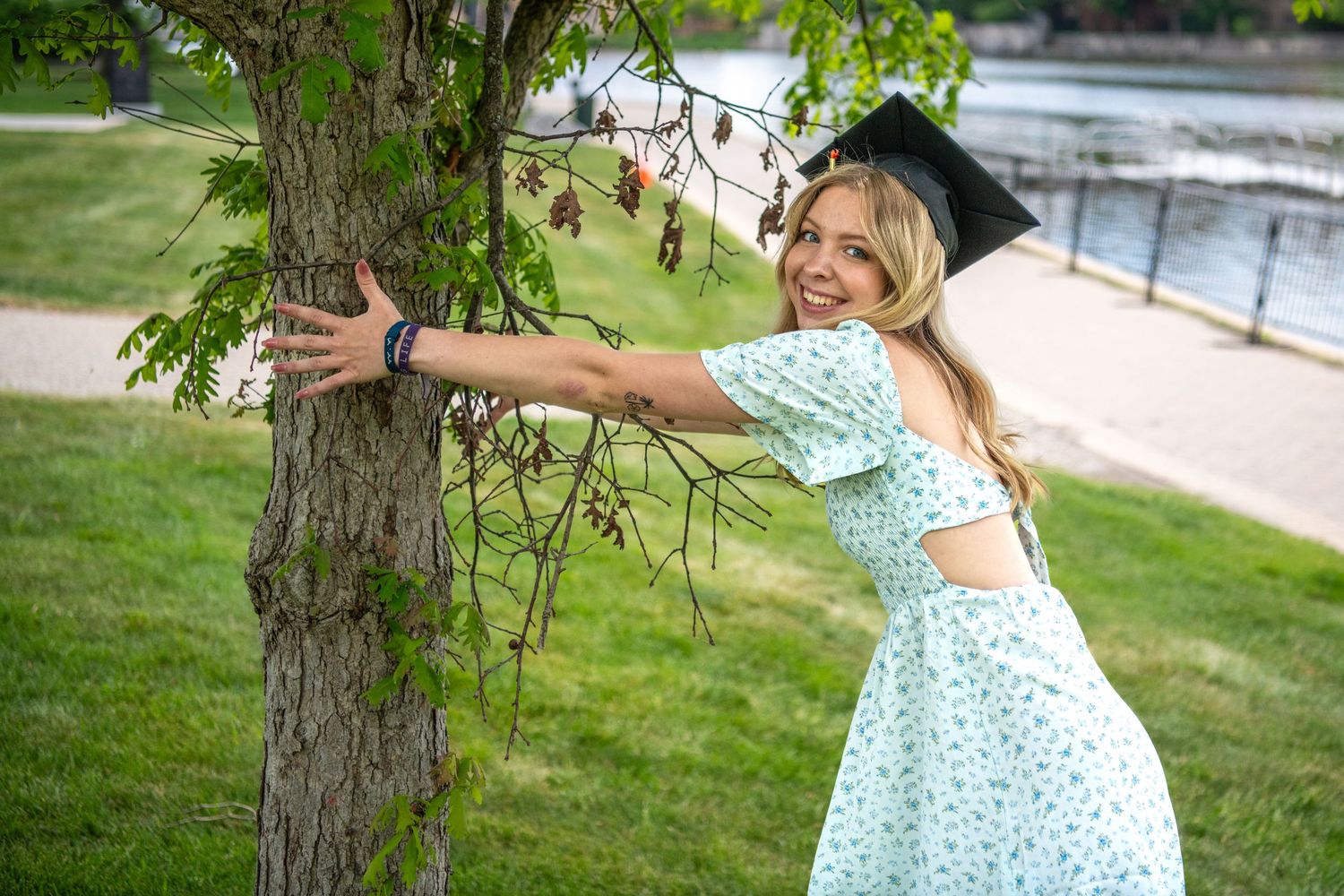 Graduate in a light blue dress poses playfully by a tree on a sunny spring day.