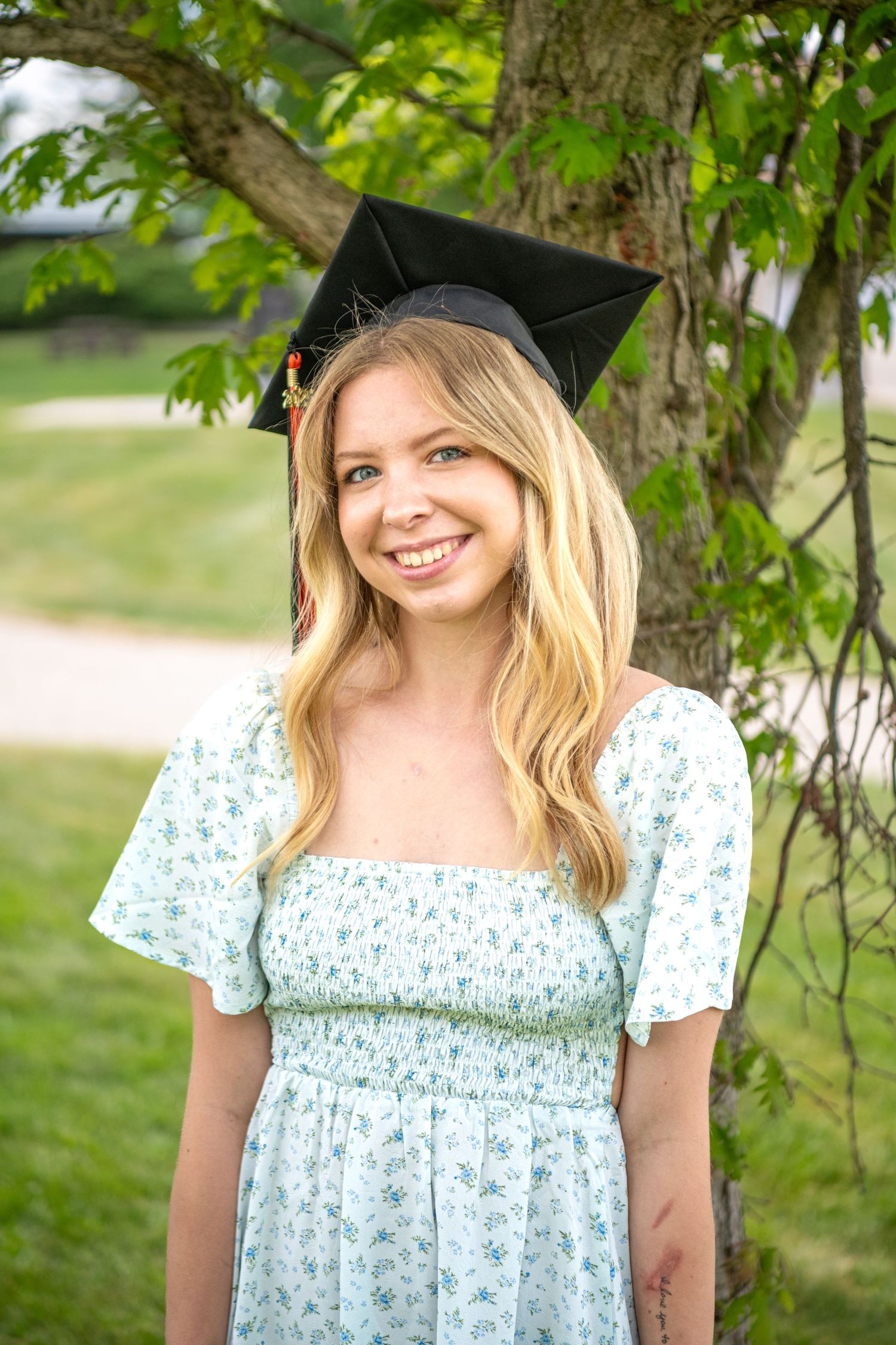Graduate in floral dress and mortarboard cap poses under green tree branches.