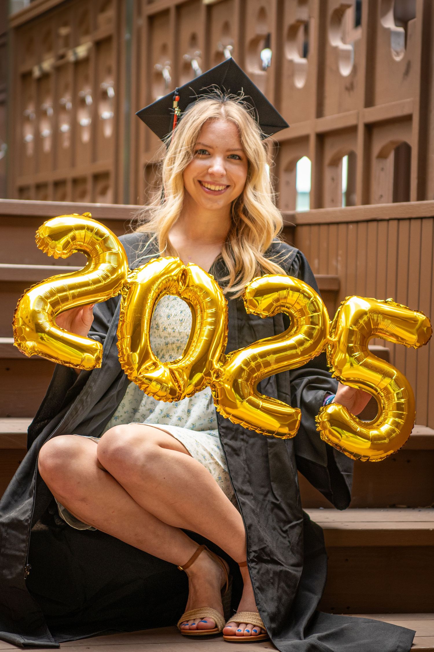 Graduate in black cap and gown poses with gold 2023 number balloons against a wooden locker background.