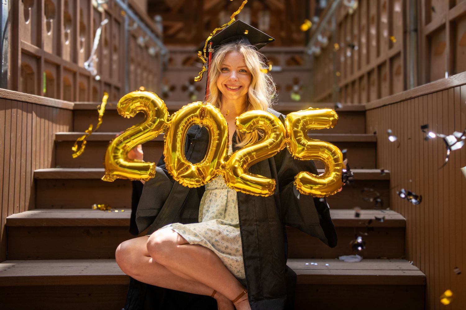 Graduate celebrating with gold letter balloons and confetti in a rustic wooden setting.