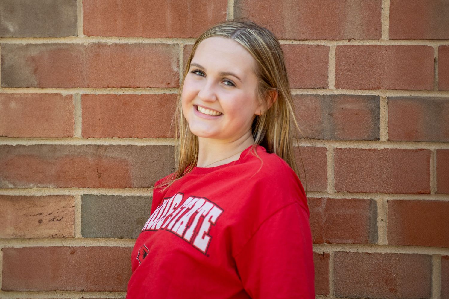 A person in a red Ohio State sweatshirt smiles while sitting against a brick wall outdoors.