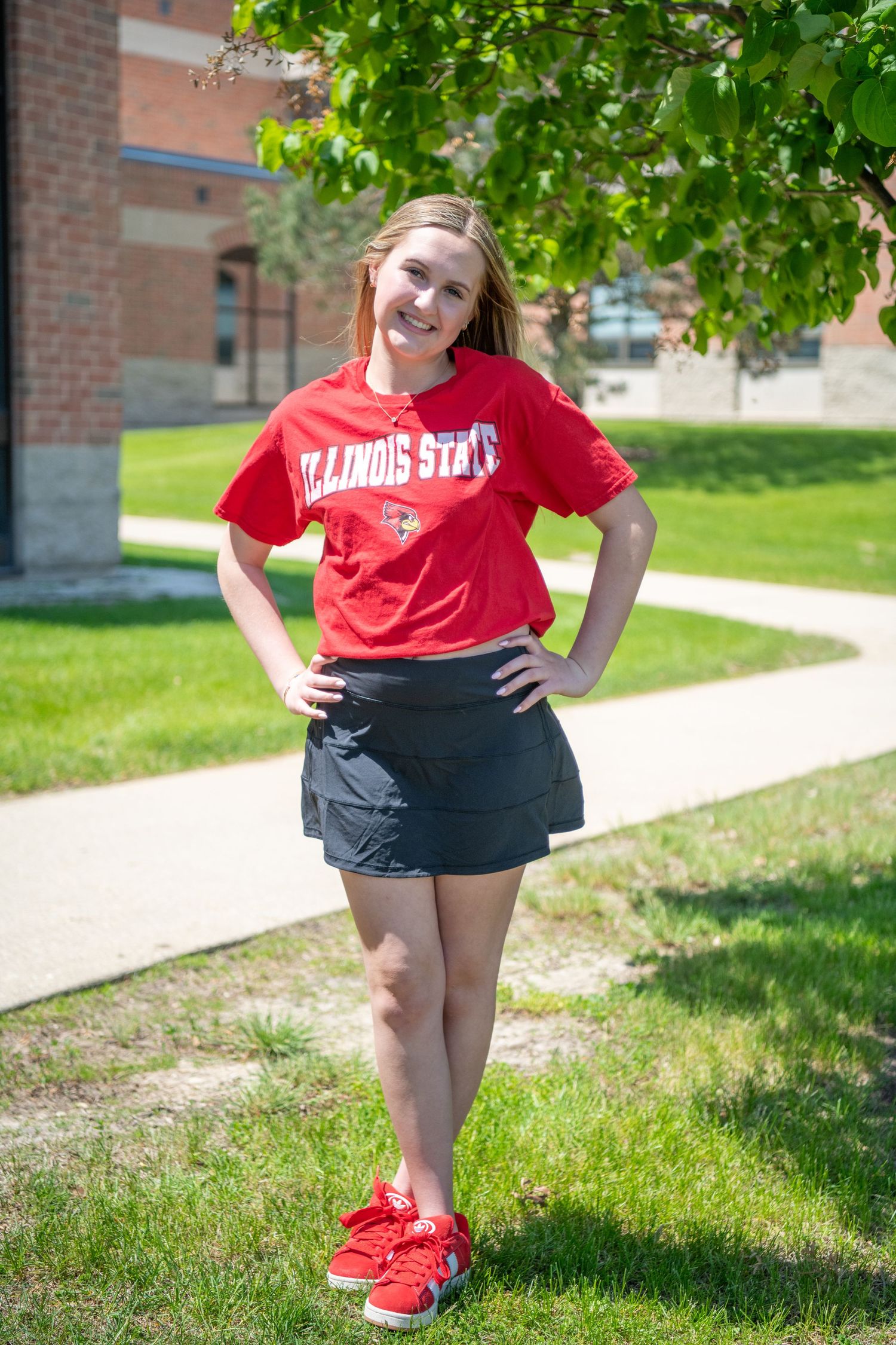 Student wearing Illinois State University red t-shirt and black skirt poses on campus sidewalk near brick building.