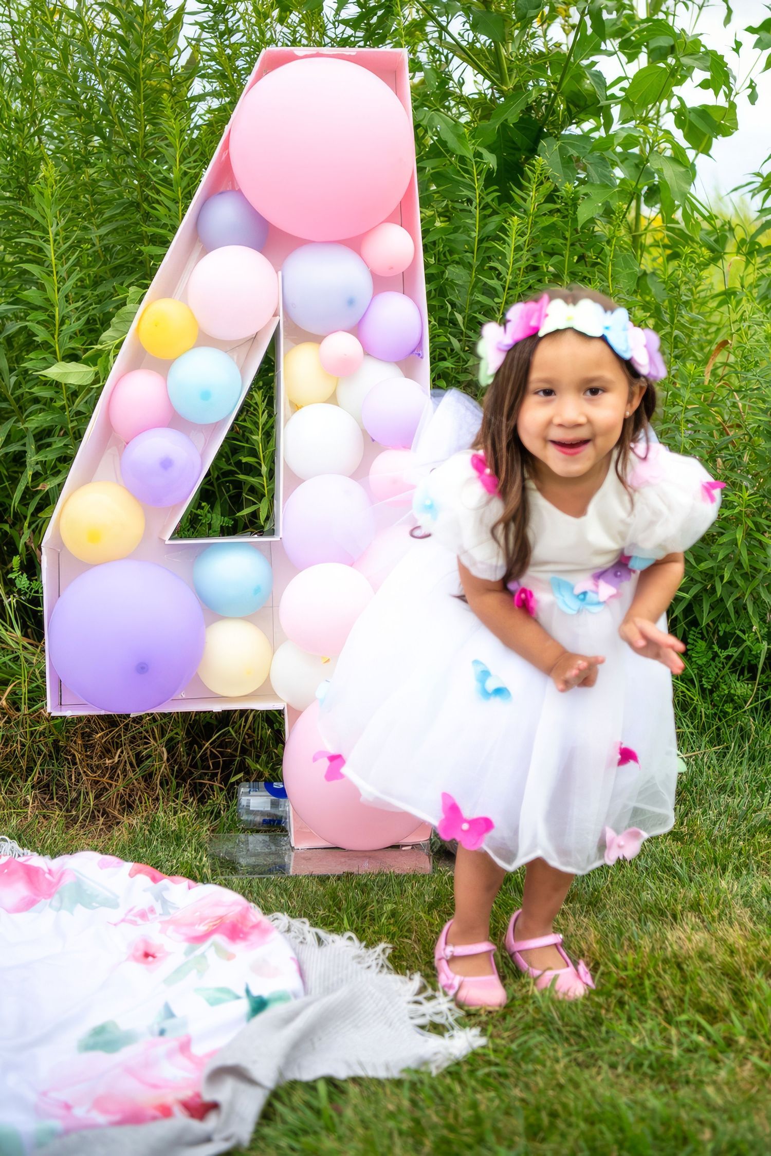 Child in white dress posing next to large balloon number 4 decorated with pastel balloons.