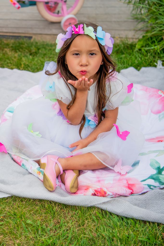 Young child in white dress with flower crown blowing kiss while sitting on blanket.