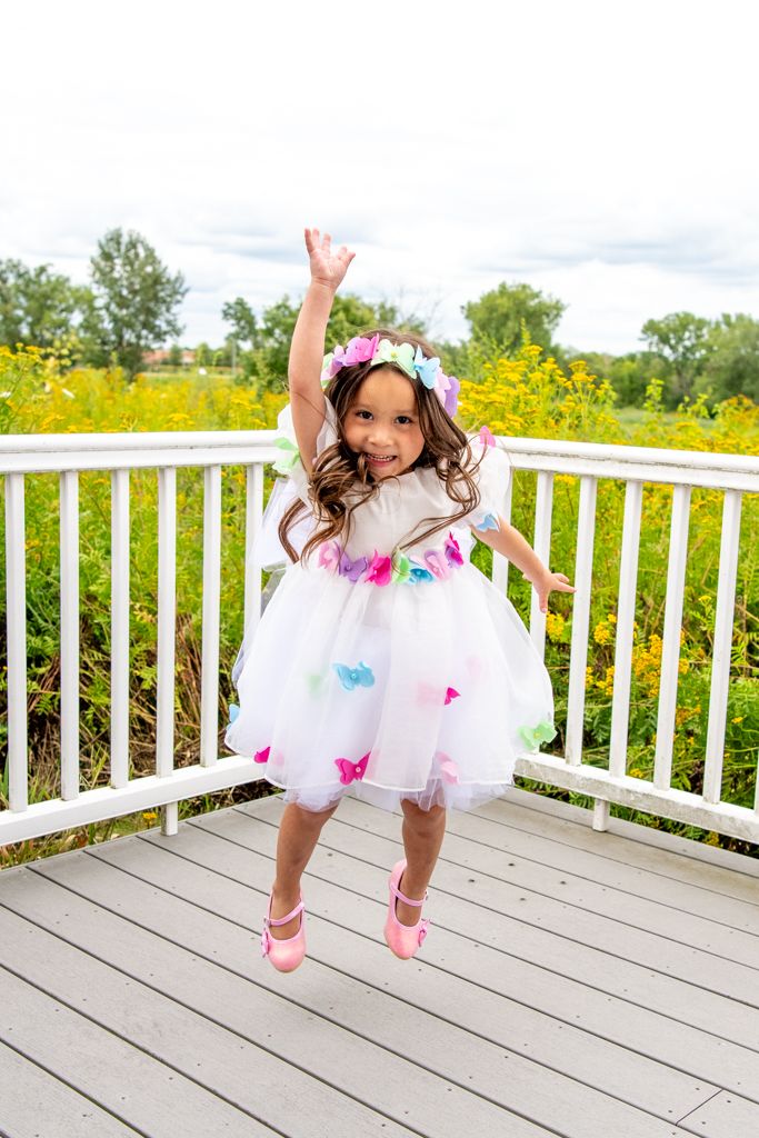 Child in white dress and floral crown jumping joyfully on wooden deck.