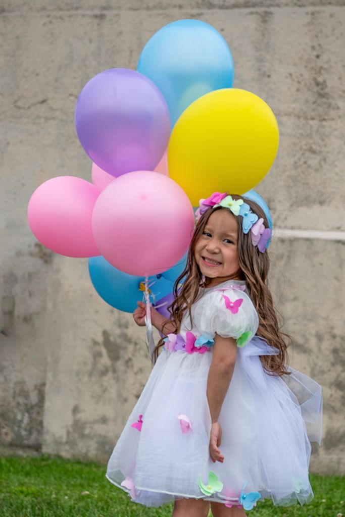 Child in white dress holding colorful balloons against concrete wall.