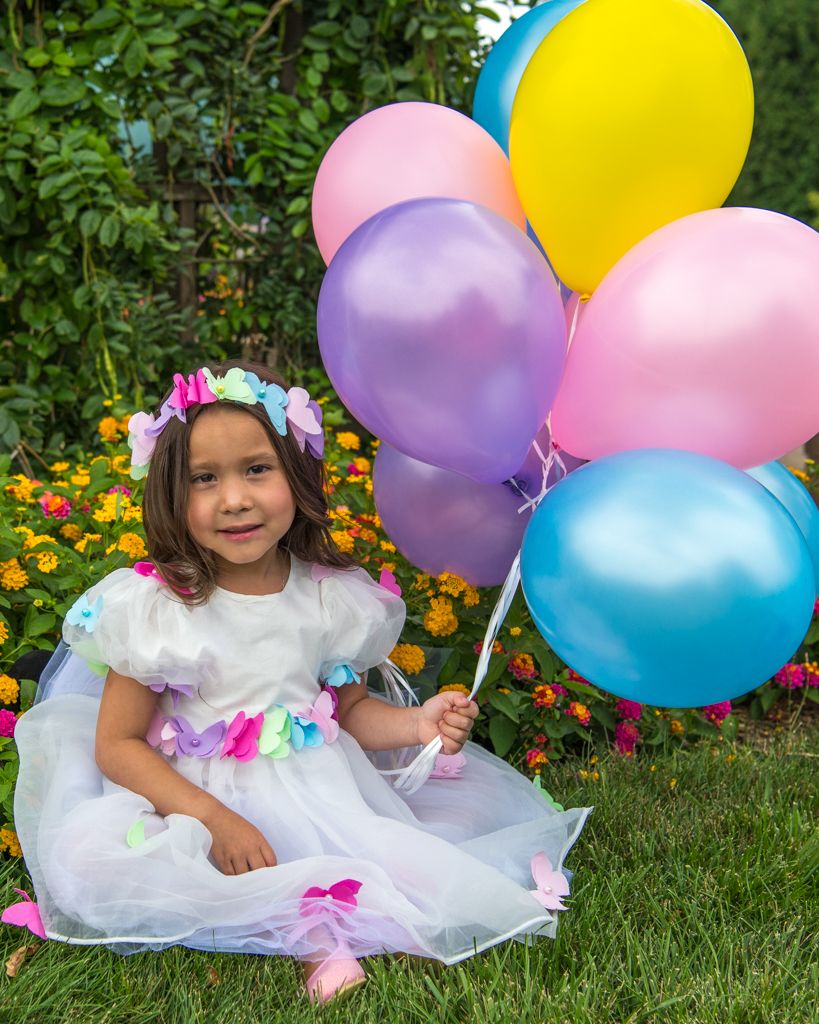 Child in floral dress sitting on grass with colorful balloons in garden setting.