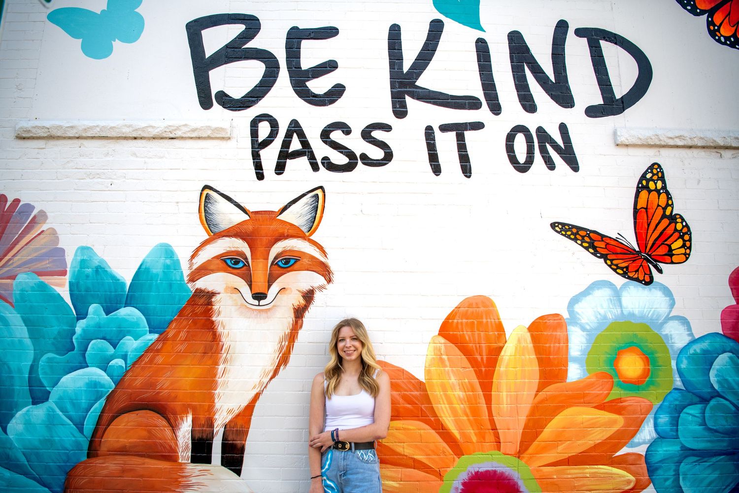 Colorful mural featuring a fox, butterflies and flowers with text reading 'Be Kind Pass It On' against white wall.
