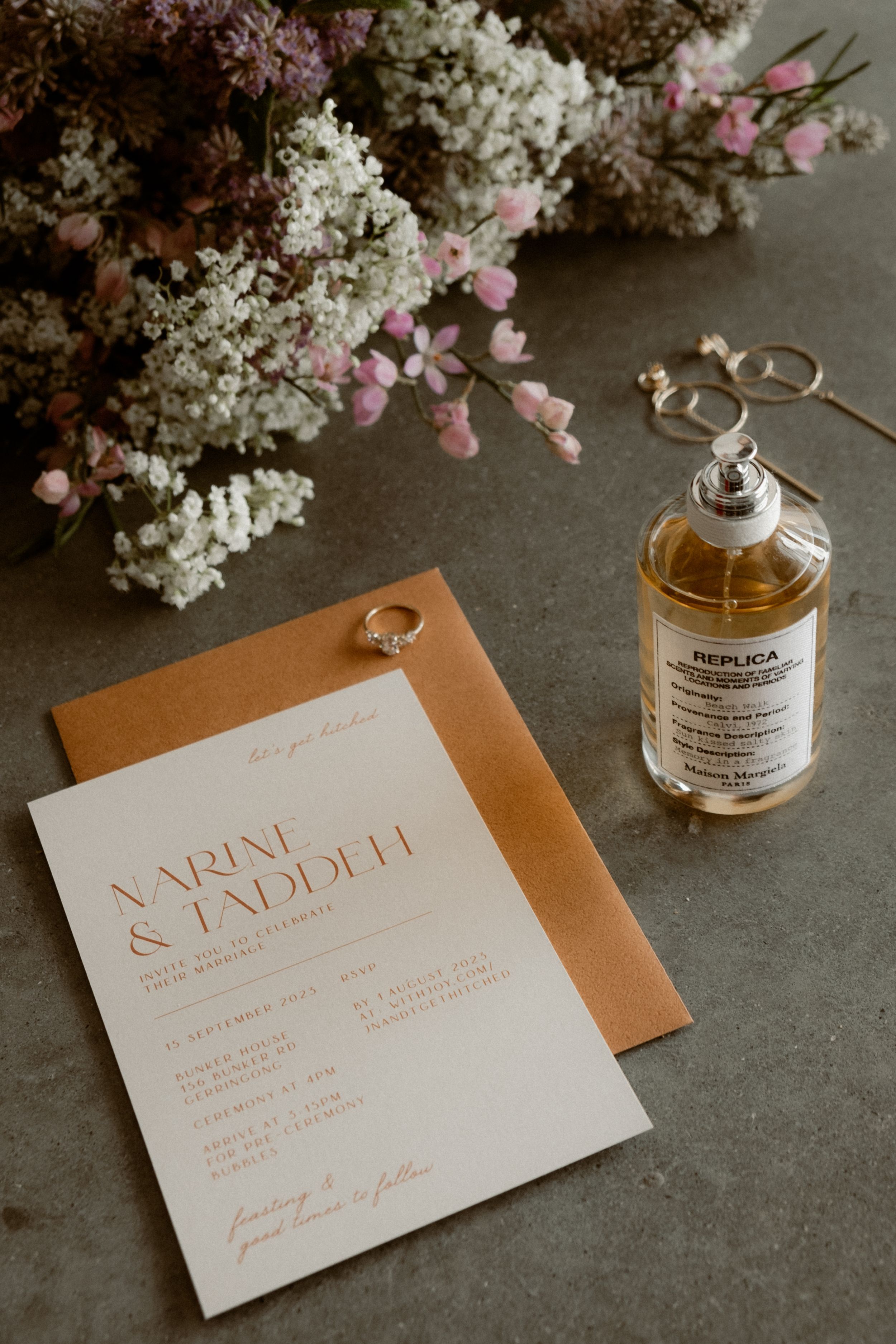 Wedding invitation with pink and white flowers, perfume bottle and rings on concrete surface.