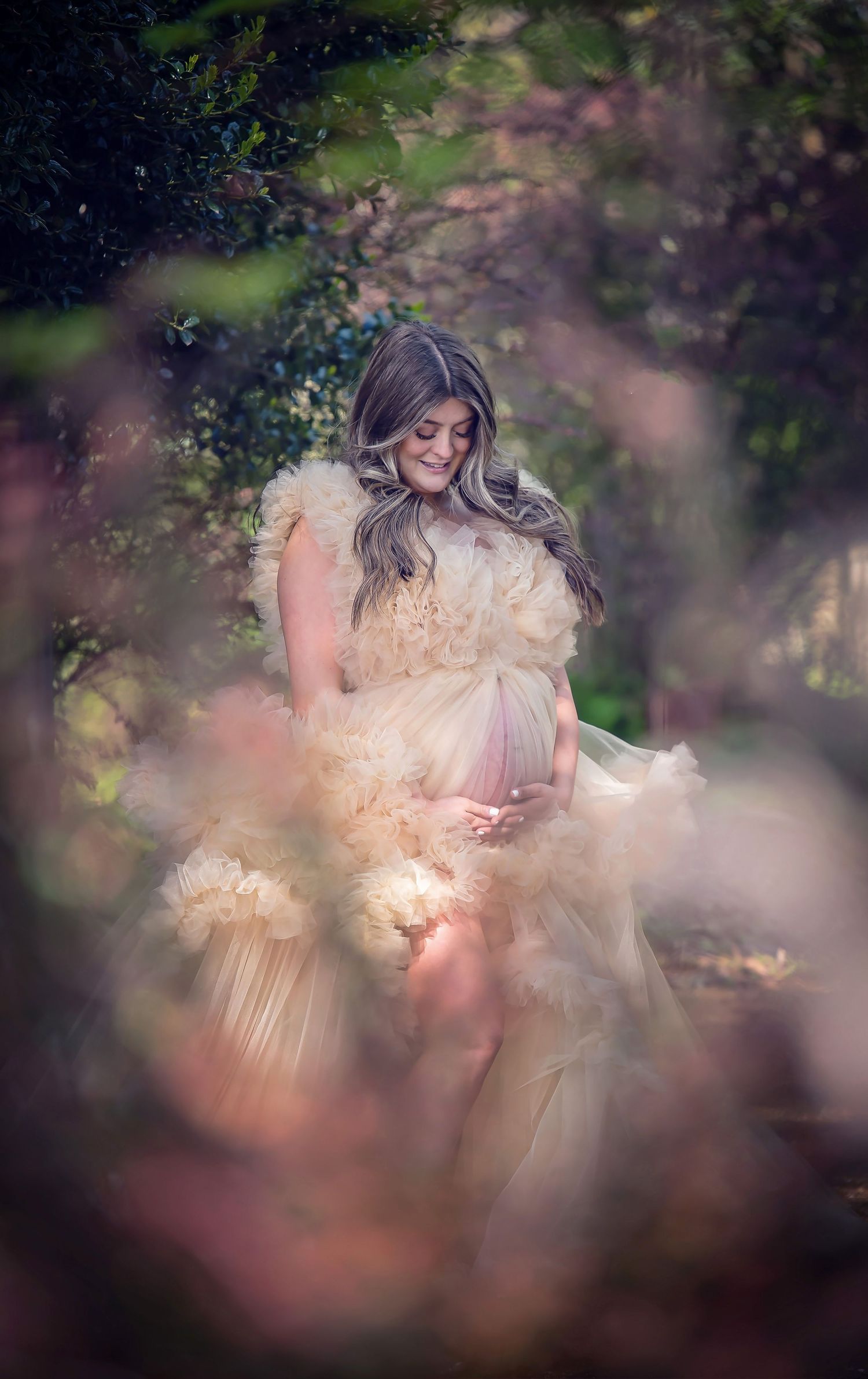 Ethereal maternity portrait of woman in flowing cream dress surrounded by dreamy bokeh and foliage.
