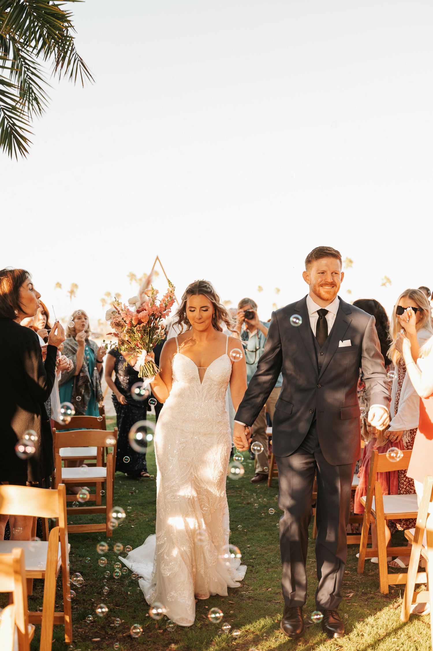 Joyful wedding recessional with palm trees and wooden chairs lining outdoor aisle.