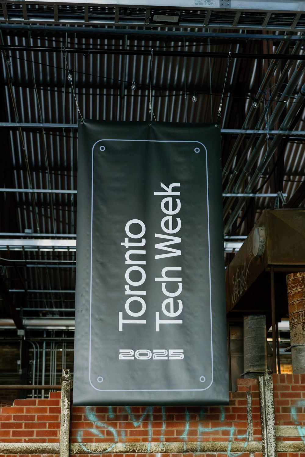 Black Toronto Tech Week 2025 sign mounted on brick wall with industrial ceiling framework above.