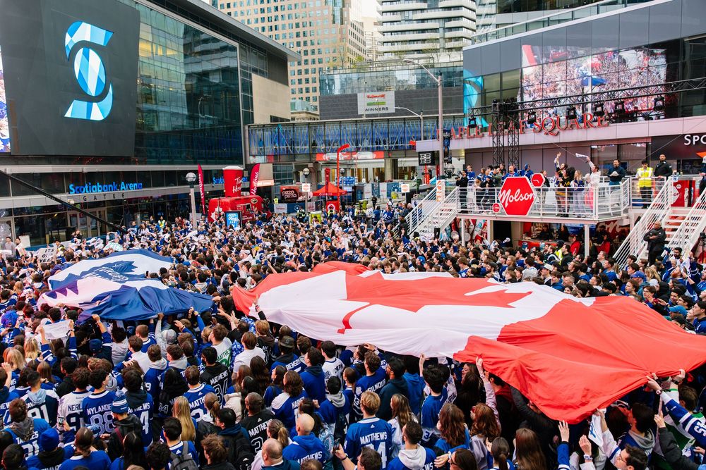 Crowd of sports fans in Toronto gather around large Canadian flag at downtown square during celebration event.