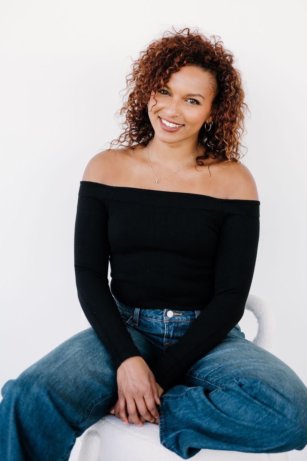 A smiling woman with curly auburn hair wearing a black off-shoulder top and blue jeans, sitting against a white background.