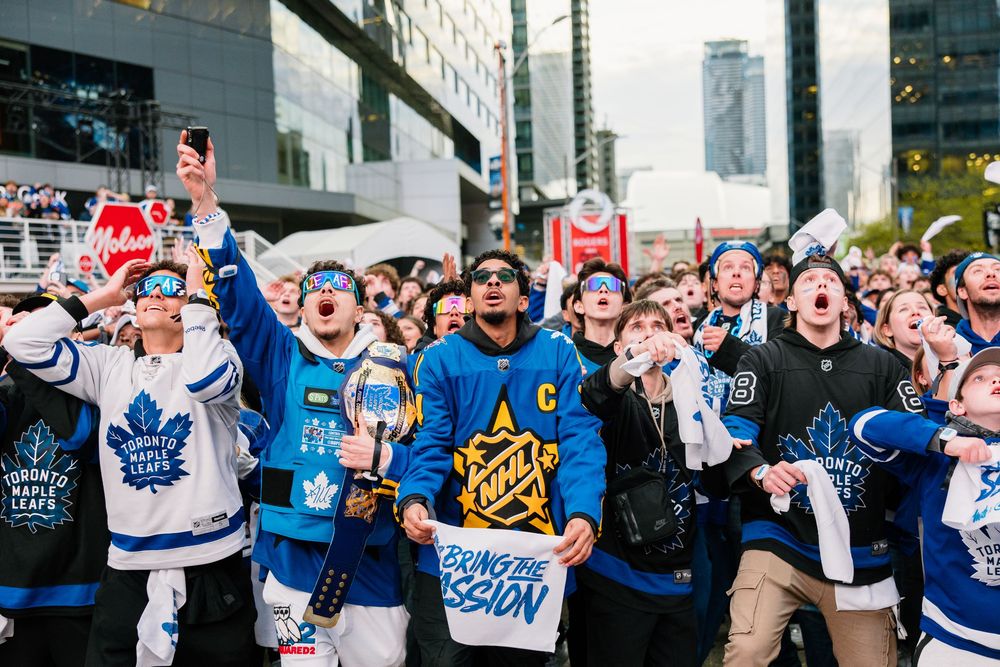 A large crowd of fans in hockey jerseys fill an outdoor plaza area waving flags and cheering at a sports event.