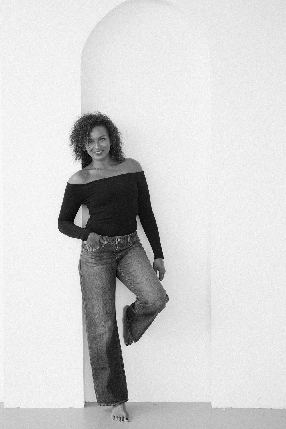 Woman in off-shoulder black top and wide-leg jeans poses against white arched wall in black and white photo.