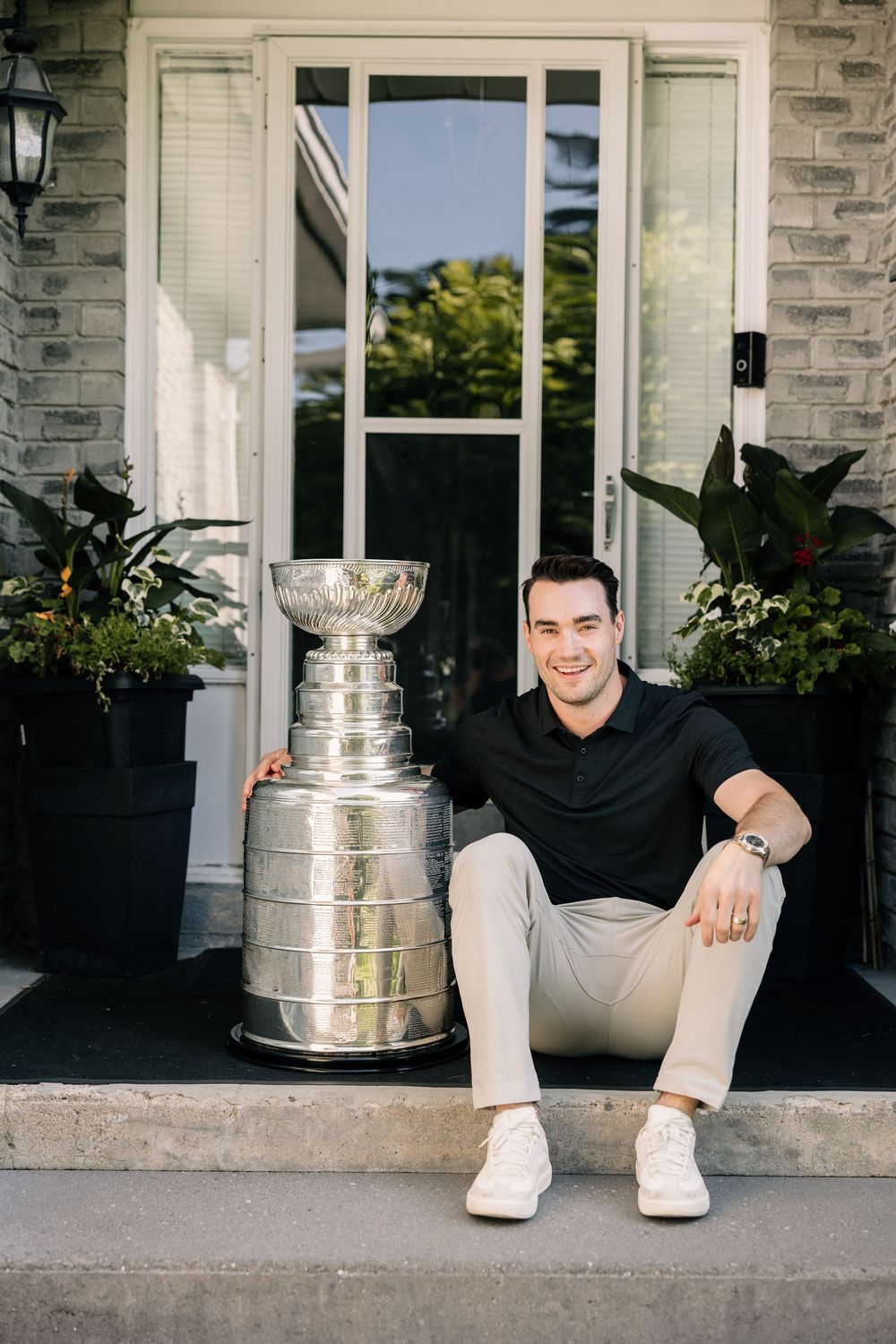 Young man in black polo and khaki pants sitting on front porch steps beside the Stanley Cup trophy.