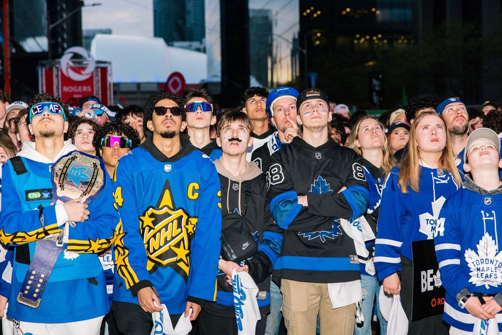 Hockey fans wearing various blue jerseys gather together to watch at an outdoor viewing event in the evening.