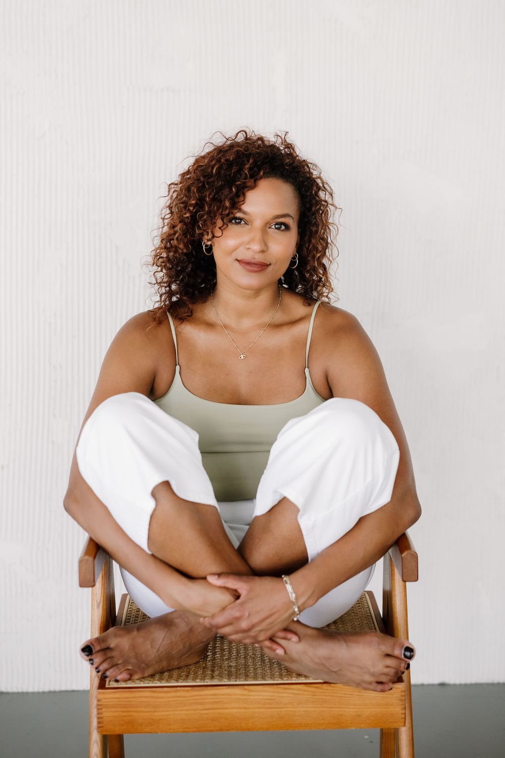 Woman with curly hair sits cross-legged on wooden chair, wearing sage green top and white pants, smiling at camera.