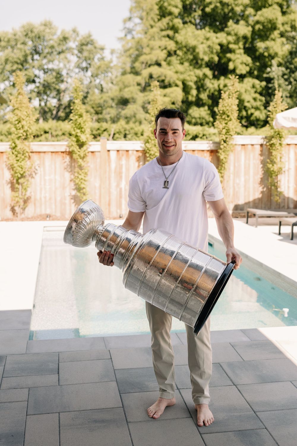 Smiling man holding the Stanley Cup trophy poolside in a white t-shirt and khaki pants on a sunny day.