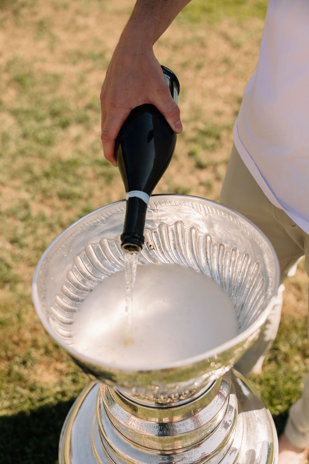 Champagne being poured into a large silver trophy cup on a grass field during a celebration.