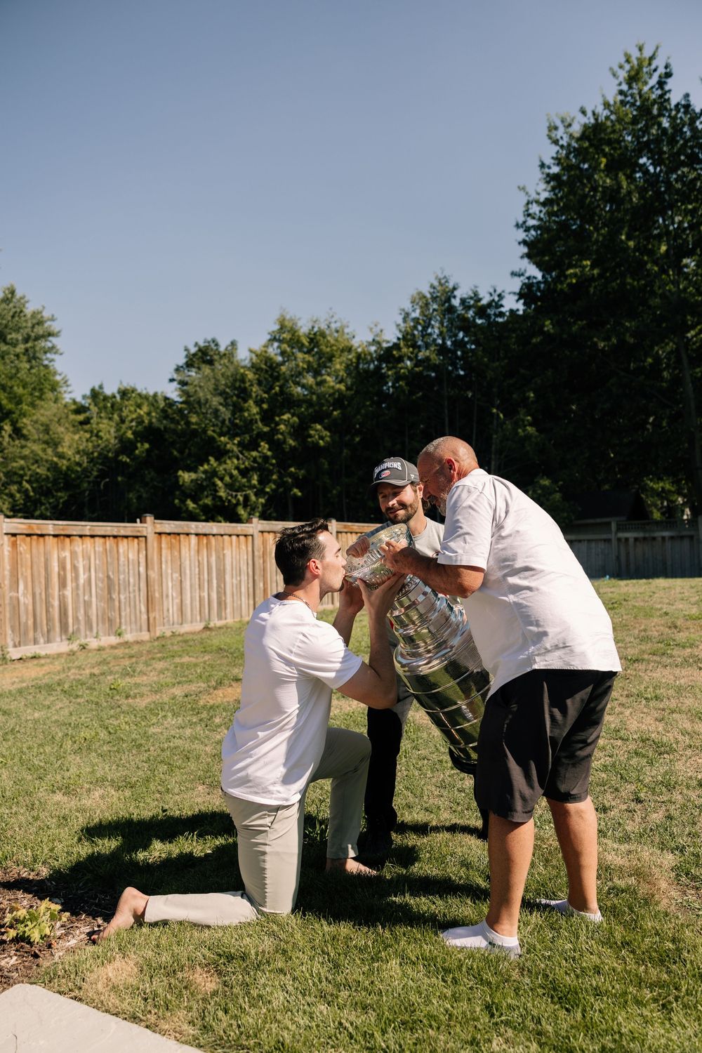 Three men in white shirts celebrate outdoors in a sunny backyard, holding a trophy together near a wooden fence.