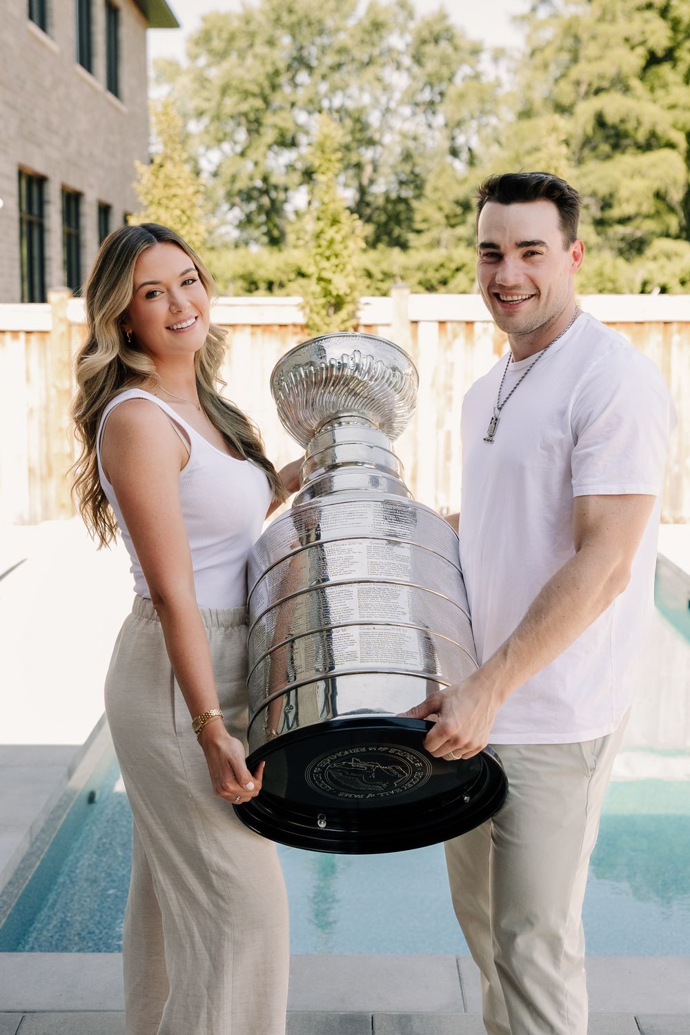 A smiling couple poses outdoors holding the Stanley Cup trophy near a pool on a sunny day.