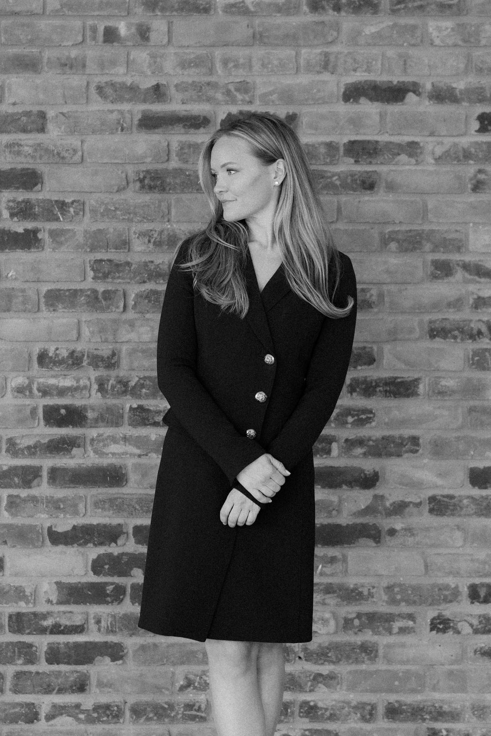 Black and white photo of a blonde woman in a dark button-front dress posing against a brick wall.