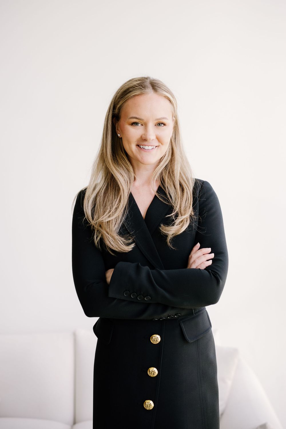 Professional woman in black outfit with gold buttons, arms crossed, smiling confidently against white background.