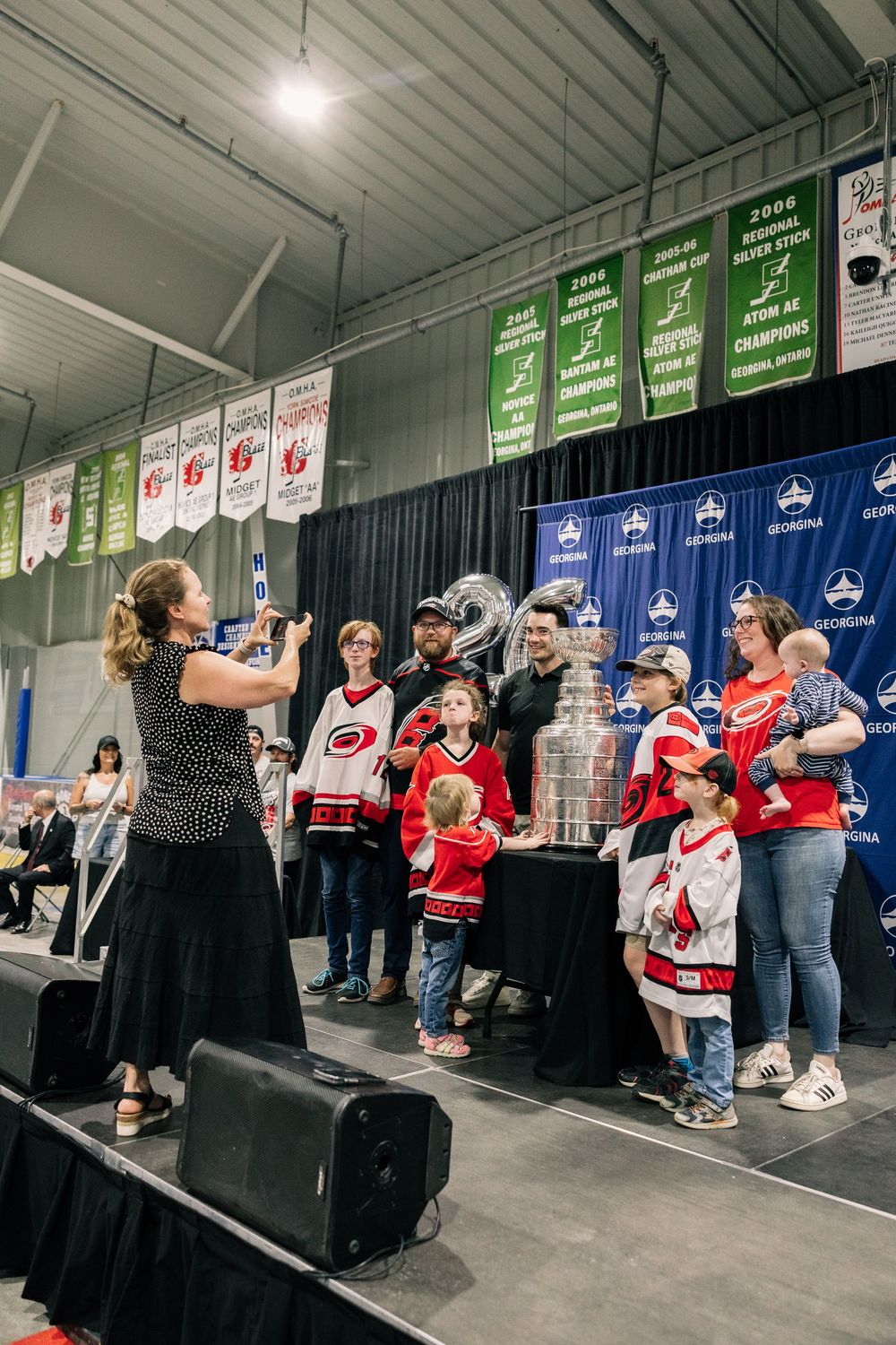 Families pose with the Stanley Cup trophy at a hockey arena event, wearing Carolina Hurricanes jerseys.