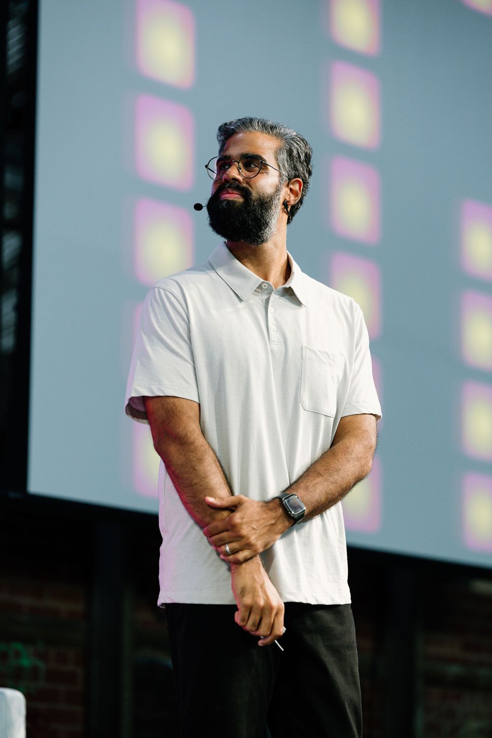 A person wearing a white polo shirt stands on stage at an event with lighting effects in the background.