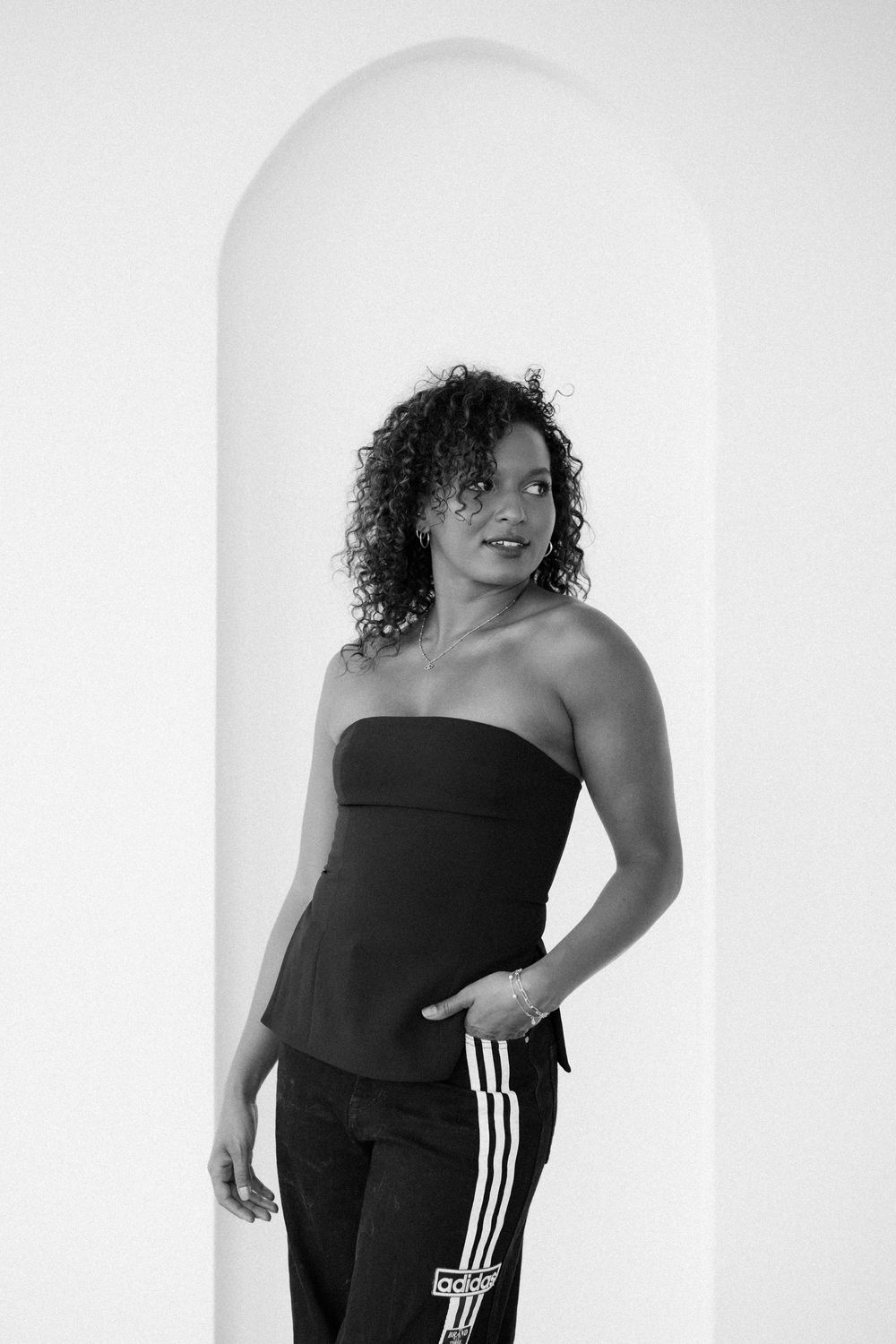 Black and white portrait of a woman in strapless top and Adidas track pants, posing in front of an arched doorway.