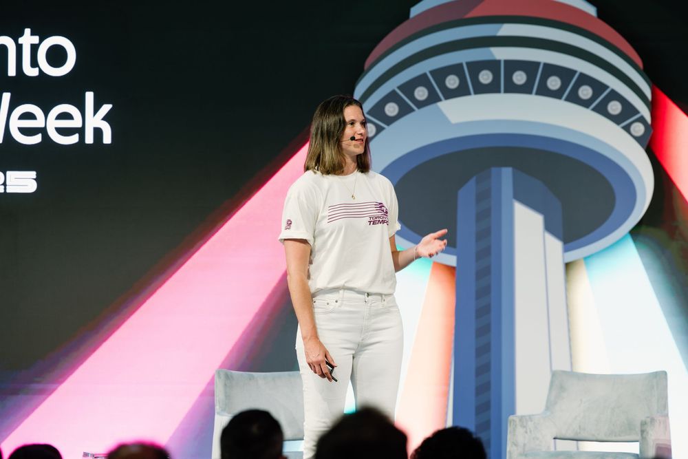 A smiling presenter in white attire stands on stage at a tech event with a futuristic tower backdrop.