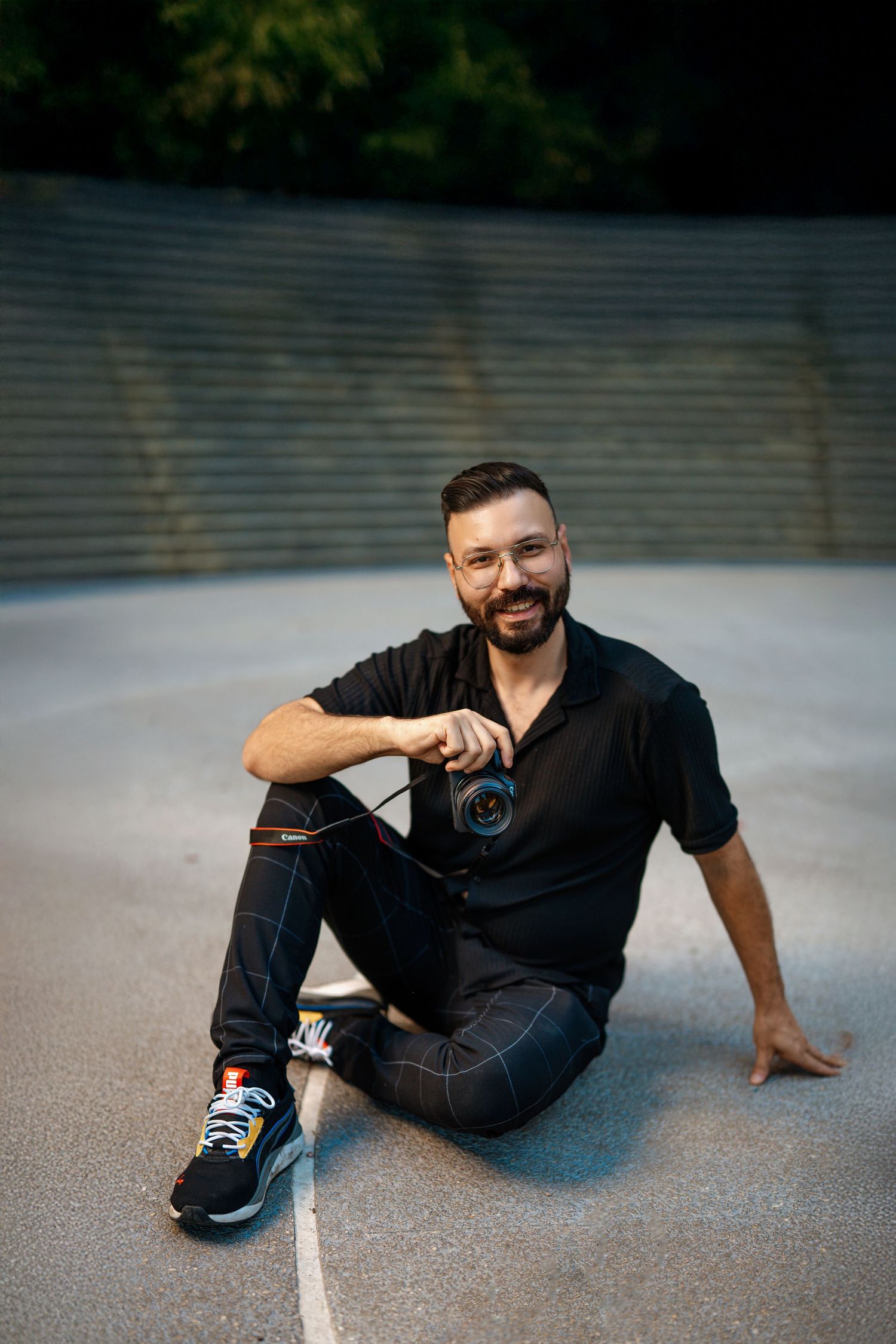 A person in a black shirt and jeans sits casually on the ground outdoors at dusk wearing athletic sneakers.