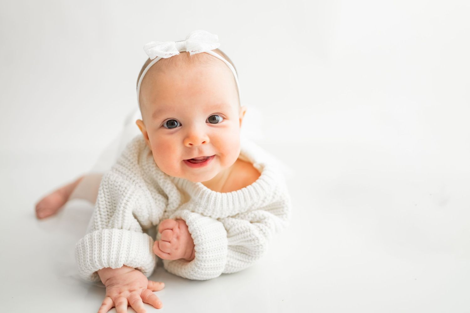 Sweet baby wearing cozy white sweater lying on white background smiling at camera.