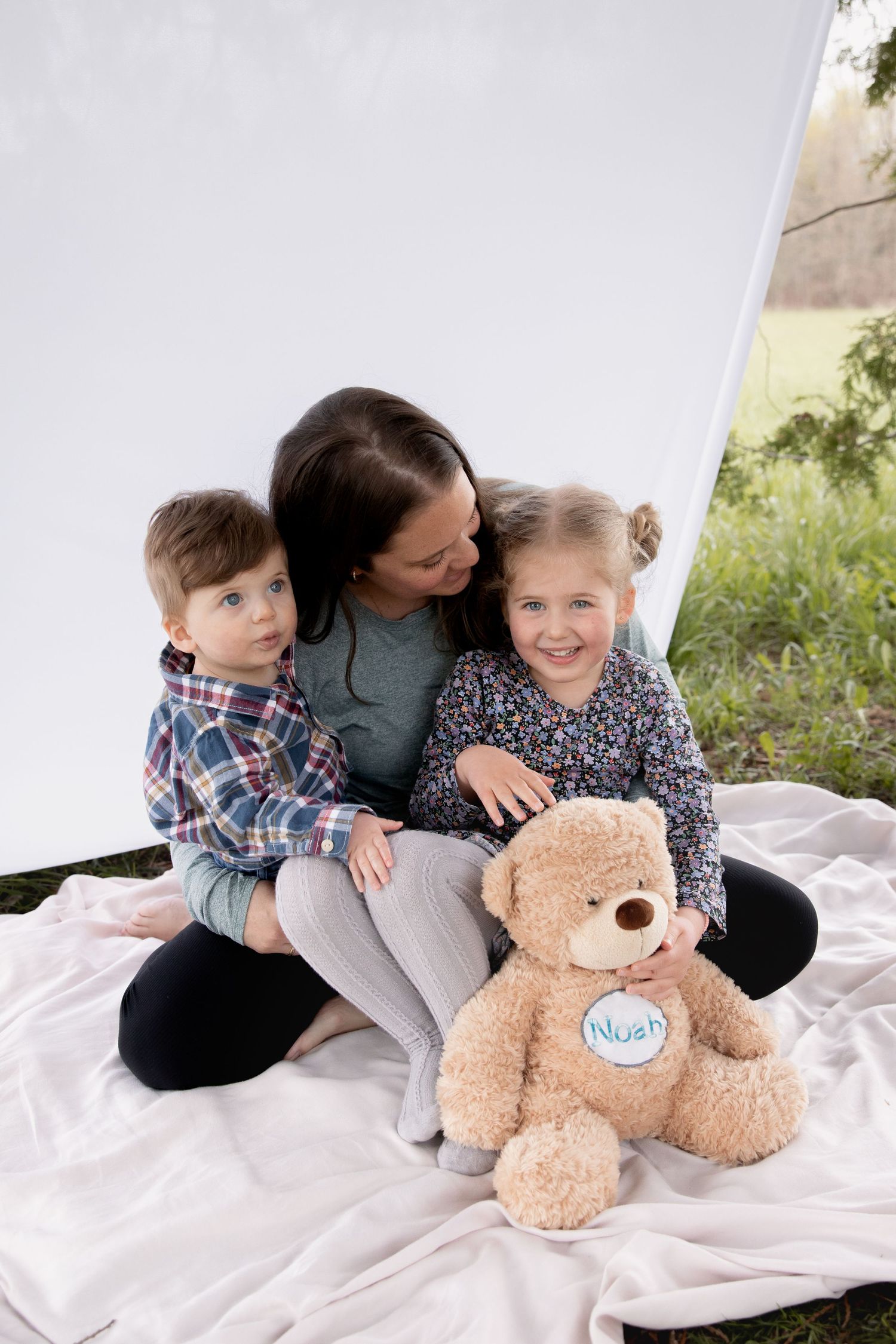 Family portrait outdoors on white blanket with plush teddy bear sitting together in casual clothing.